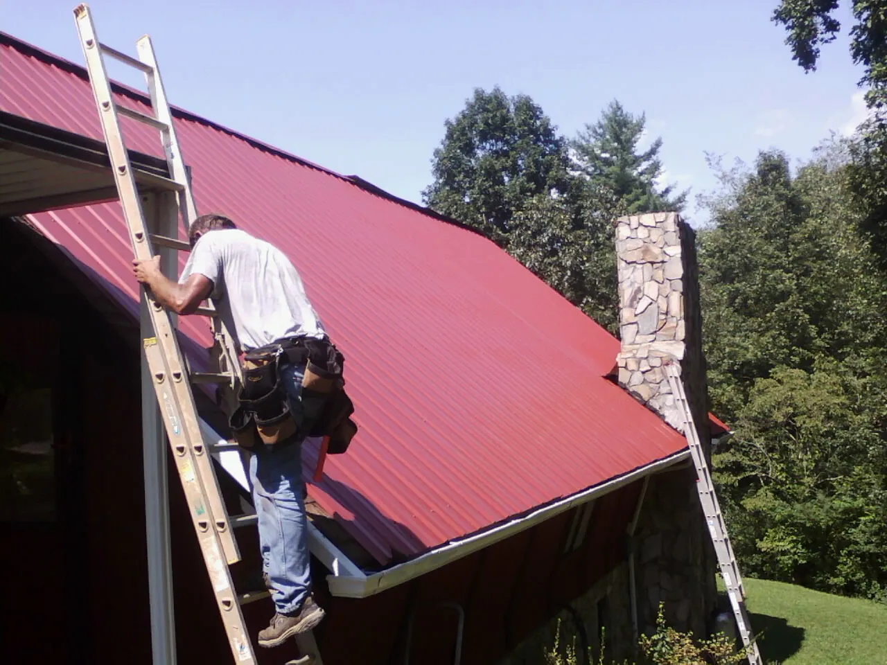 Roofer on a ladder beside a red metal roof and stone chimney — Soffit & Fascia Repair by Roofing Brothers in Snellville